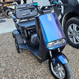 Blue electric scooter on a gravel surface with a vehicle in the background