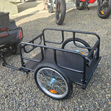 Black bicycle cargo trailer on a gravel surface with a motorcycle in the background.