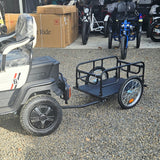 Small trailer attached to a golf cart on a gravel surface with other vehicles and boxes in the background.