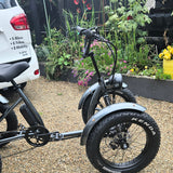 Four-wheeled electric bike with 'Kenda' tires on a gravel surface, with a white vehicle and garden in the background.