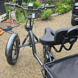 Black electric quad bike with a seat on a gravel surface with plants and a car in the background