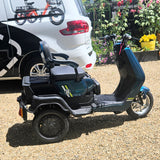 Electric scooter with a child seat parked on a gravel surface, with a vehicle and plants in the background.