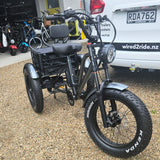 Black electric tricycle with large wheels on a gravel surface, with a white van labeled 'wired2ride.nz' in the background.